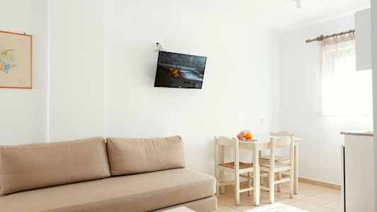 Ett ljust rum med beige soffa, vägghängd TV, vit matgrupp med stolar och ett fönster med ljusa gardiner. (AI-genererad text)