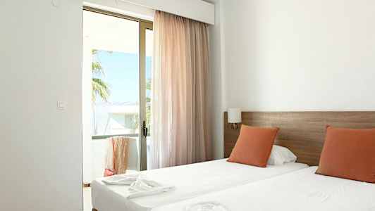 Ett hotellrum med två enkelsängar, orangea kuddar och en glasdörr som leder till en balkong. (AI-genererad text)