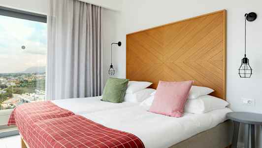 Hotellrum med två bäddar, färgglada kuddar, rödplädar och modernt inrett, med utsikt genom stora fönster. (AI-genererad text)