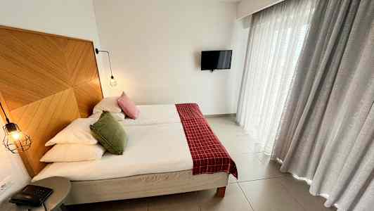 Bild av ett hotellrum med en dubbelsäng, en träpanel som sänggavel, röda och gröna prydnadskuddar, upphängda lampor, en liten bordstelefon och gardiner. En TV hänger på väggen. (AI-genererad text)