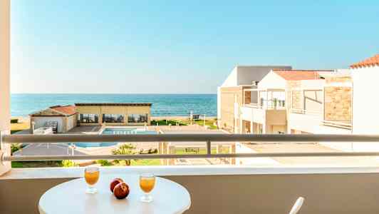 Utsikt från en balkong med bord där två glas och två äpplen står, mot en pool, byggnader och havet. (AI-genererad text)