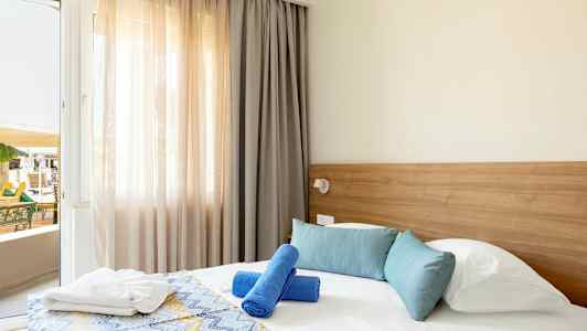 Ett hotellrum med en säng som har blå och vita kuddar samt två blå badhanddukar. En fönsterdörr är öppen mot en balkong. (AI-genererad text)