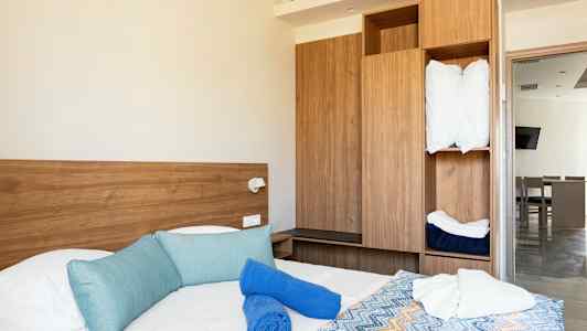 Ett hotellrum med en säng som har blå kuddar, blå och vita handdukar samt ett färgglatt mönstrat överkast. I bakgrunden syns en träfärgad garderob och en dörr mot ett annat rum. (AI-genererad text)