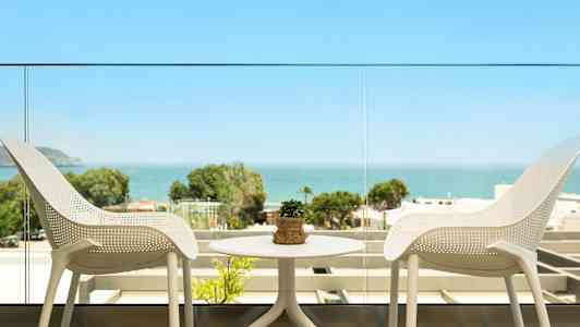 Utsikt från en balkong med två vita stolar och ett bord med en liten växt, samt havet och grönska i bakgrunden. (AI-genererad text)