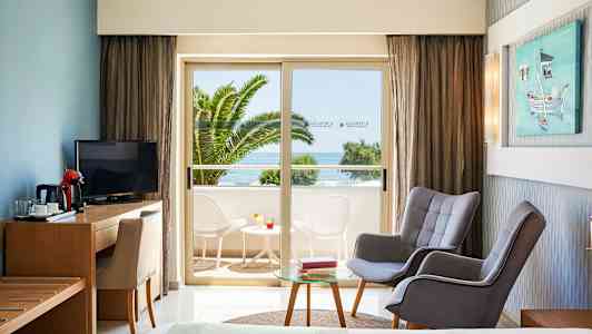 Hotellrum med sittgrupp och utsikt mot balkong med bord och stolar samt havsutsikt. (AI-genererad text)