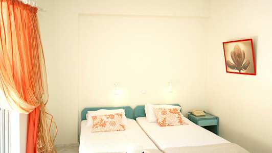 Två enkelsängar med orange mönstrade kuddar, en telefon på ett blått nattduksbord och orange gardiner vid fönstret. (AI-genererad text)