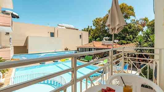 Utsikt från en balkong mot en pool och hotellområde med solstolar, parasoller och grönska. På balkongen finns ett bord med en tallrik melon och ett glas juice. (AI-genererad text)