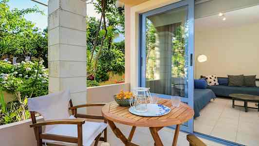 En utomhusterrass med ett träbord och stolar, omgiven av gröna växter. En glasdörr leder till ett rum med sittmöbler. (AI-genererad text)