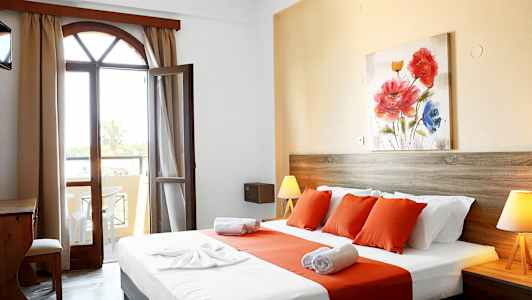 Ett hotellrum med en säng klädd i vita och orange lakan, ett träskrivbord och en öppning mot en balkong. (AI-genererad text)