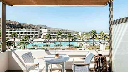 Utsikt från en balkong med ett bord och två stolar, som visar en pool, palmer och moderna byggnader i bakgrunden. (AI-genererad text)