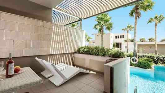 En utomhusterrass med två vita solstolar och ett bord med vin och frukt. Framför terrassen finns en privat pool och grön vegetation. (AI-genererad text)