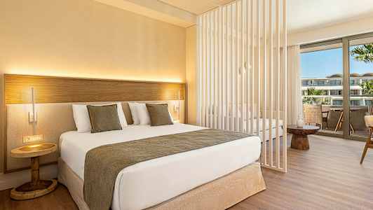 Ett modernt hotellrum med en dubbelsäng, dekorativa kuddar och en liten nattduksbord. Rummet har en skiljevägg och en sittgrupp med utsikt över en balkong. (AI-genererad text)