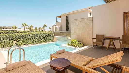 Bild föreställer en terrass med solstolar, ett litet bord och två stolar bredvid en privat pool. I bakgrunden syns grönskande buskar och palmer. (AI-genererad text)