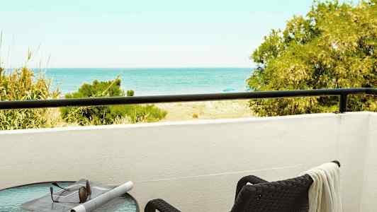 En balkong med utsikt mot havet, ett bord med solglasögon och en tidning samt en stol med en handduk. (AI-genererad text)