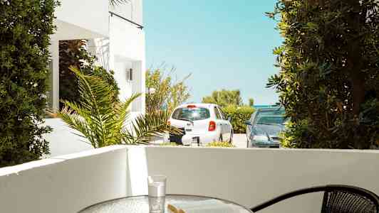 Terrass med ett bord där en bok och ett glas står. Utanför syns parkerade bilar och grönska. (AI-genererad text)