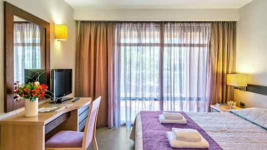 Ett hotellrum med en säng klädd i lila överkast, skrivbord med en TV och blombukett, samt ett stort fönster med genomskinliga gardiner. (AI-genererad text)