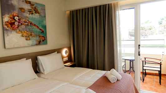 Ett hotellrum med två enkelsängar, en konstmålning på väggen och glasdörrar som leder till en balkong med bord och stolar. (AI-genererad text)