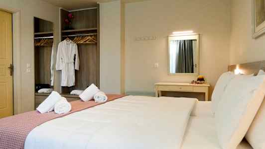 Ett hotellrum med en dubbelsäng, sängkläder i vitt och en rosa filt. En garderob med hängande klärob och spegel syns i bakgrunden. (AI-genererad text)