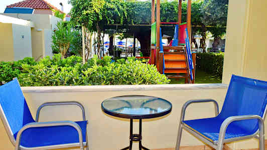 Två blå stolar vid ett bord med glasbordsskiva på en terrass, med lekplats och grönska i bakgrunden. (AI-genererad text)