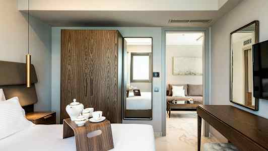 Ett hotellrum med säng, tebricka, garderob och spegel. I bakgrunden syns en sällskapsdel med soffa. (AI-genererad text)