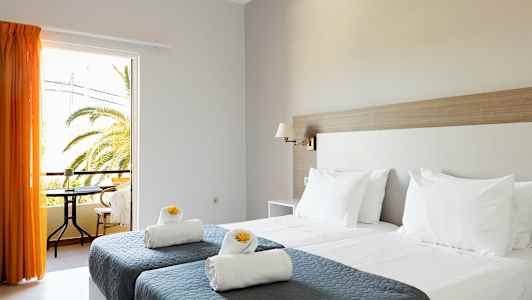 Ett hotellrum med två enkelsängar med grå överkast och vita kuddar samt handdukar dekorerade med gula blommor. Ett fönster med orange gardiner öppnar mot en balkong med utsikt över palmer. (AI-genererad text)