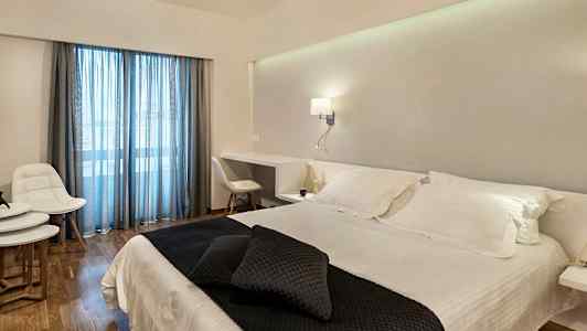 Ett hotellrum med en dubbelsäng klädd i vita lakan och mörka prydnadskuddar. Rummet har ett skrivbord, vita möbler och glasdörrar med blåa gardiner. (AI-genererad text)