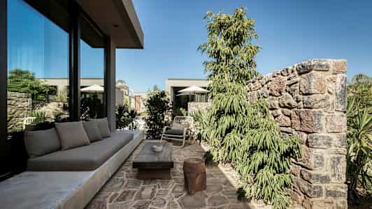 Stenbelagd uteplats med soffmöbler, bord och stolar nära ett glashus, omgiven av växter och murar. (AI-genererad text)