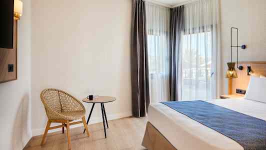 Ett hotellrum med en modern stol, ett litet bord och en säng bredvid fönster med ljusa gardiner. (AI-genererad text)