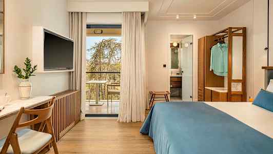 Hotellrum med dubbelsäng, skrivbord och TV. Glasdörrar leder ut till en balkong. Badrum synligt genom en öppen dörr. (AI-genererad text)