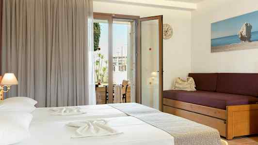 Ett hotellrum med två sängar klädda i ljusa lakan, en lila soffa med kuddar, och en glasdörr som leder ut till en balkong. (AI-genererad text)