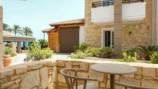 En uteplats med ett bord och stolar omgiven av stenmur, med utsikt mot en byggnad med stenfasad och palmer i bakgrunden. (AI-genererad text)