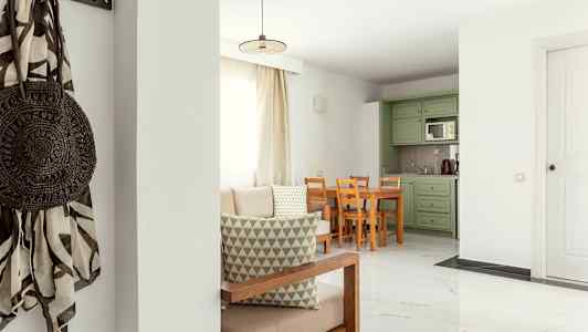 Ett vardagsrum med ljus inredning, bord och stolar samt gröna köksskåp. (AI-genererad text)