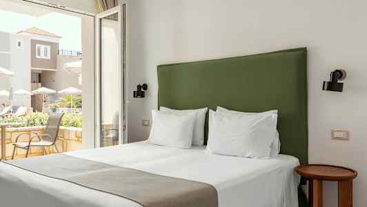 Hotellrum med en säng och ett grönt sänggavel, ljusa väggar och fönster med utsikt över en terrass. (AI-genererad text)