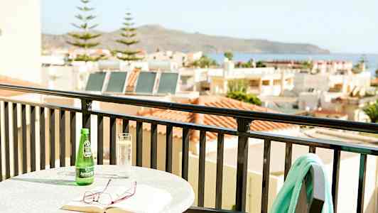 Bord med bok, glasögon och dryck på balkong med utsikt över hus och hav. (AI-genererad text)