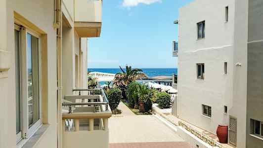 Byggnader med balkonger öppnar mot en promenad kantad av palmer, med havet i bakgrunden. (AI-genererad text)