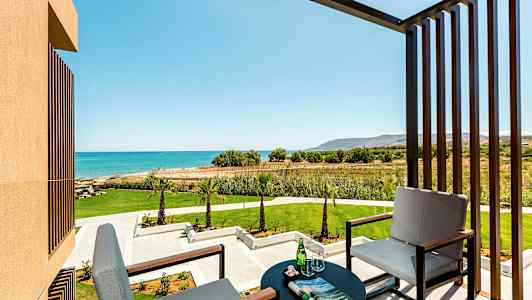 Bild av en balkong med två sittplatser, ett bord med glas och tidning, med utsikt mot en grön trädgård och blått hav. (AI-genererad text)