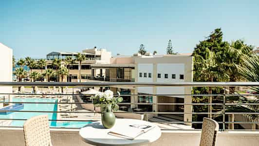 Utsikt från en balkong med ett bord, en stol och en vas med blommor, samt en öppen bok. I bakgrunden syns en byggnad och solområde med pool. (AI-genererad text)