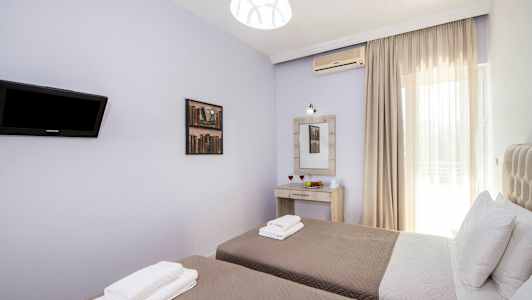 Bild av ett rum med två enkelsängar, sängkläder och kuddar, en väggmonterad TV, en spegel med belysning ovanför ett skrivbord och en fönster med ljusa gardiner. (AI-genererad text)