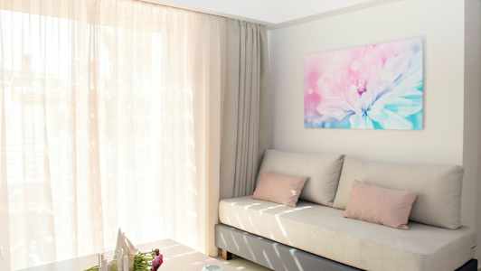 Ett ljust och luftigt vardagsrum med en beige soffa, rosa kuddar och ett stort fönster med tunna gardiner. På väggen hänger en blomsterbild. (AI-genererad text)