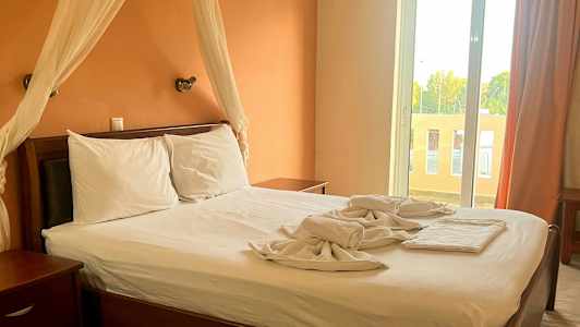 Ett sovrum med en dubbelsäng, vita lakan och en orange vägg. Ett stort fönster med gardiner syns i bakgrunden. (AI-genererad text)