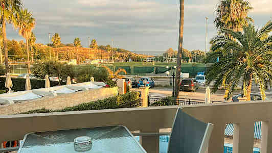 Utsikt från en terrass mot en idrottsplats omgivet av palmer och parkeringsplatser. (AI-genererad text)