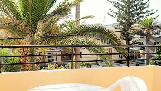 Vy från balkong med ett bord och plaststol, palmträd och byggnader i bakgrunden. (AI-genererad text)