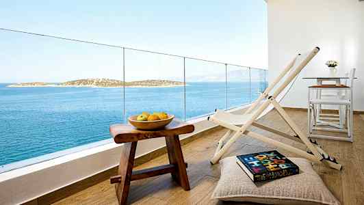 Utsikt från en balkong med stolar, ett bord, och en skål med frukt på ett träbord med havet och öar i bakgrunden. (AI-genererad text)