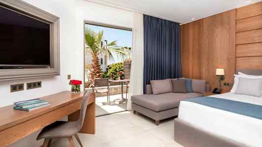 Ett hotellrum med en bädd, en soffa, ett skrivbord och en stor TV. Utsikt mot en terrass med träd och möbler. (AI-genererad text)