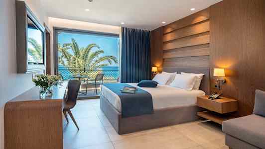 Ett hotellrum med säng, nattduksbord, skrivbord och stora glasdörrar som leder till en balkong med utsikt över palmer och havet. (AI-genererad text)