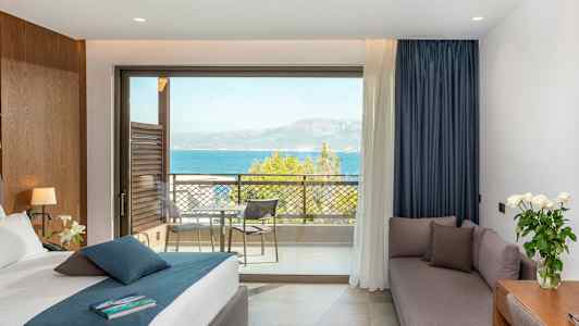 Hotellrum med säng, soffa, skrivbord och utsikt över havet från en balkong. (AI-genererad text)