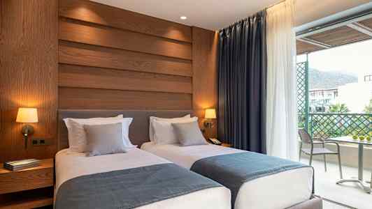 Hotellrum med två enkelsängar, dekorerade med blå överkast och grå kuddar, vägglampor, samt utsikt mot en balkong. (AI-genererad text)