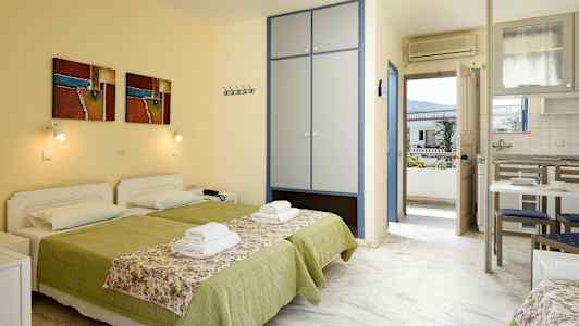 Två bäddar med grön sängklädsel och blommönstrade överkast i ett rum med skåp, köksbord och dörr till balkong. (AI-genererad text)