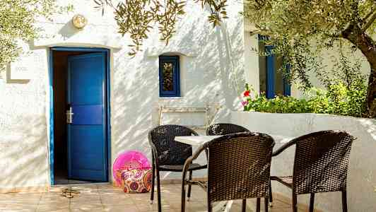 Vit byggnad med blå dörr och fönster, bord och stolar på terass. (AI-genererad text)