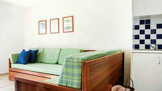 En träsoffa med gröna rutiga dynor, två blå kuddar och tre inramade bilder på väggen. Del av ett kök ses med rutigt kakel. (AI-genererad text)